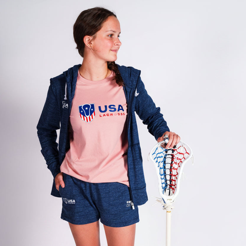 A woman modeling a pink USA Lacrosse graphic t-shirt, navy blue zip-up hoodie, and matching navy blue shorts, holding a lacrosse stick against a light gray studio background.