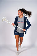A woman modeling a USA Lacrosse athletic set, featuring a heather gray t-shirt with a red, white, and blue logo, a navy blue zip-up hoodie, and matching navy blue shorts, set against a plain light gray studio background.