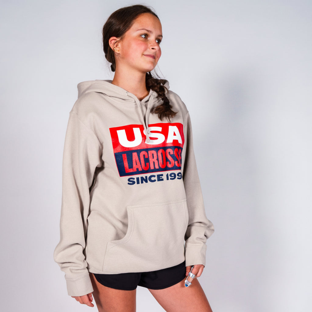 A woman modeling a light beige hoodie with a red and blue 'USA LACROSY SINCE 1998' graphic on the chest, paired with black athletic shorts, against a light gray studio background.